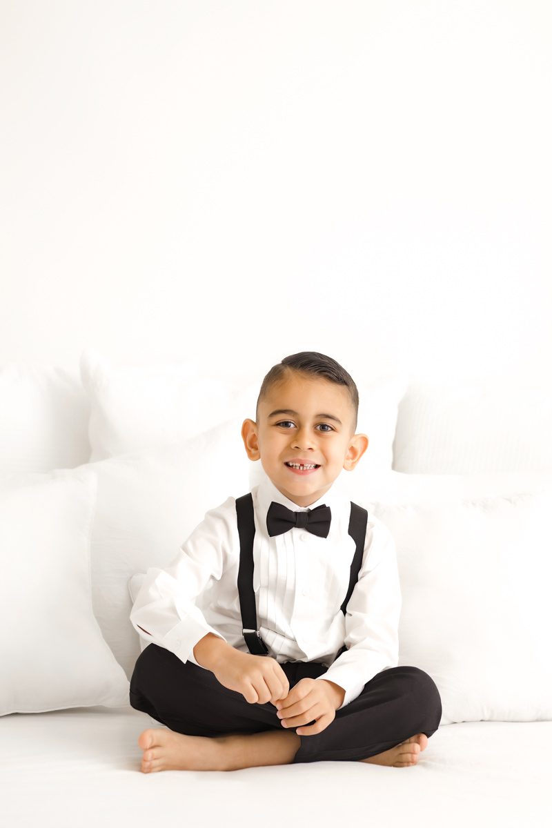 A young boy in a white shirt, vest, and bow tie sits smiling against a white backdrop during a family session.