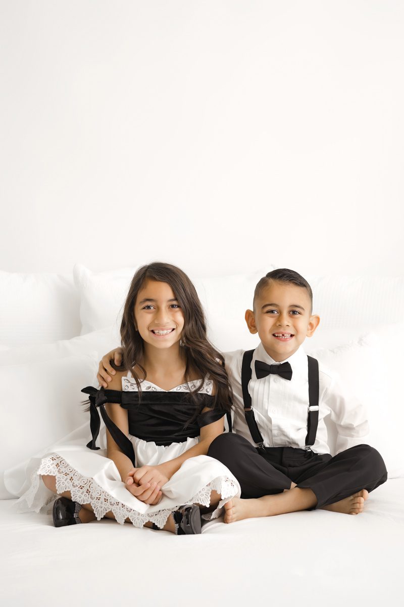 Two children dressed in formal attire sitting together on a white backdrop during a family photography session.