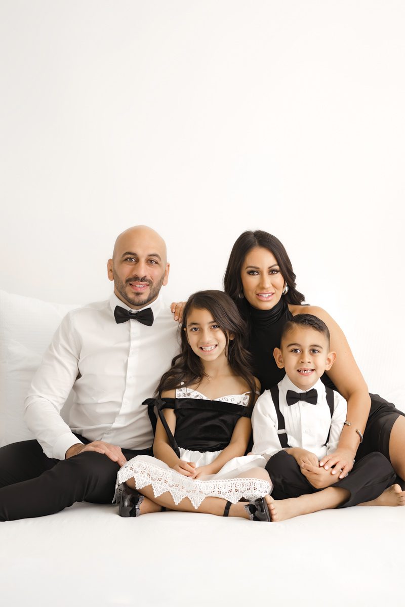 A family of four, dressed in formal black and white attire, smiling while sitting together on a white sofa during their family photoshoot.