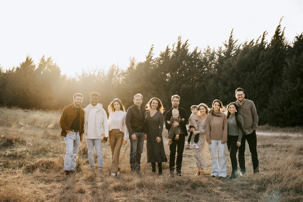 A family photoshoot featuring a group of adults, some holding children, casually walking together in a field at sunset.