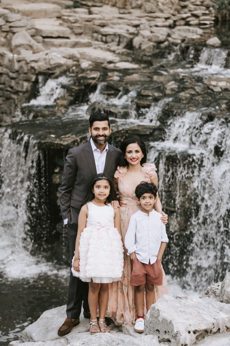 A family of four posing for a family photoshoot in front of a small waterfall, with the parents standing behind and the two children seated in front.