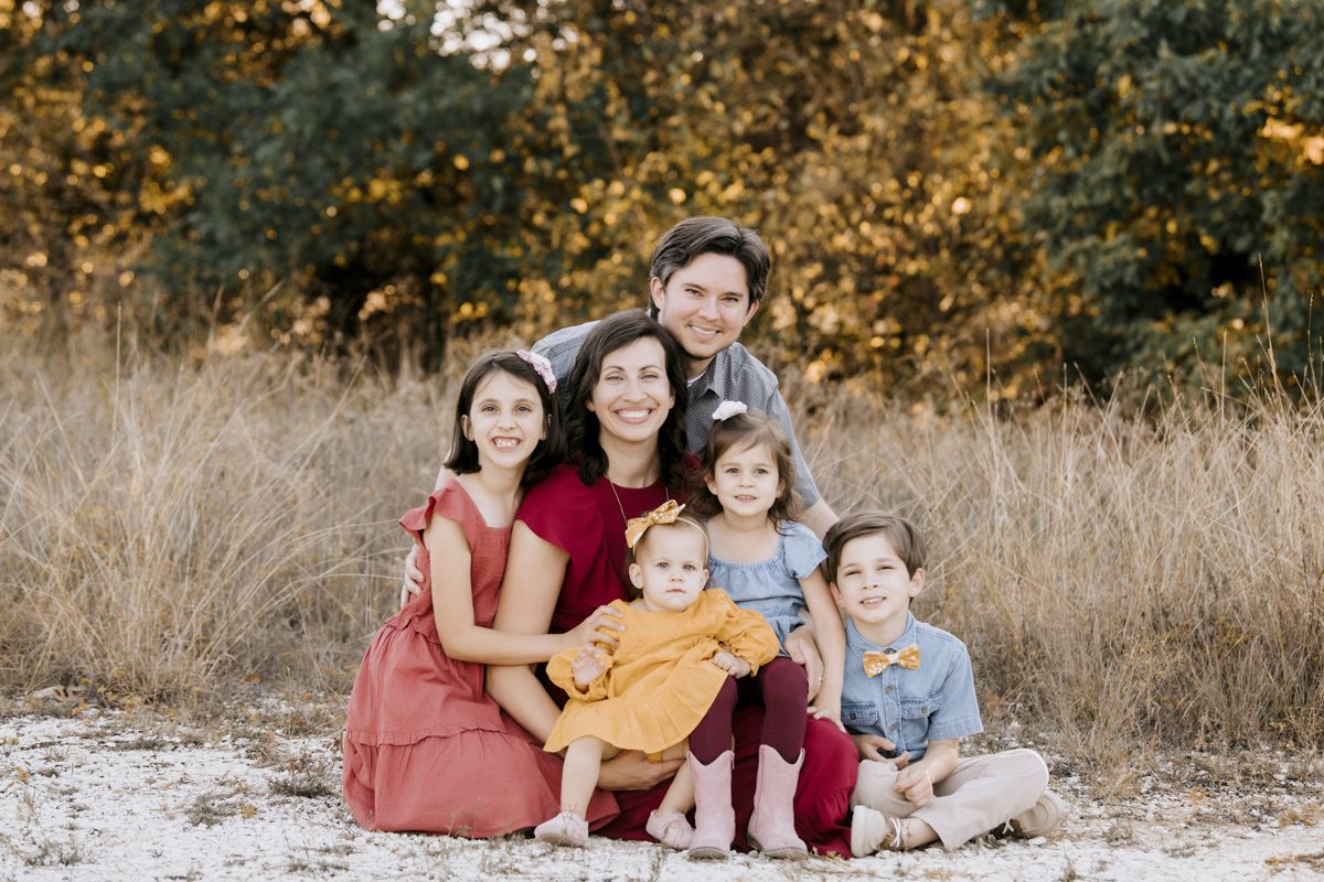 A family photoshoot with two adults and four children sitting outdoors with a backdrop of trees.