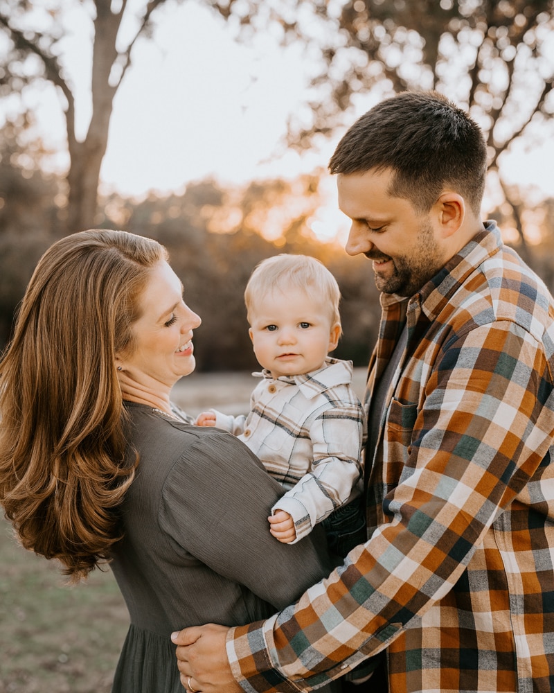 couple stands outdoors, smiling at baby for dallas family photography